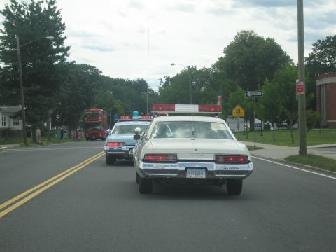 1991 Ford LTD Crown Victoria - cream-colored sedan and a marked police cruiser drive down a tree-lined residential street, a red double-decker bus ahead.