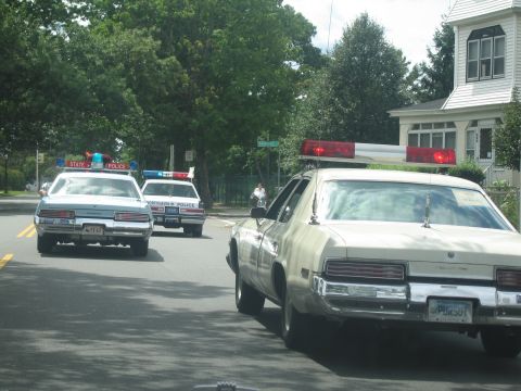 1991 Ford LTD Crown Victoria - cream Buick with lightbar and several State Police cruisers travel along a street past a white house at a parade.