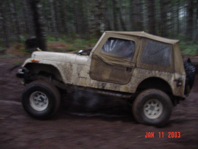 Mud-covered tan Jeep CJ with soft top mid-motion on a dark forest trail