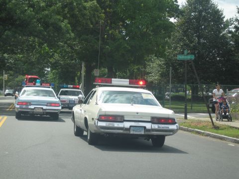 1991 Ford LTD Crown Victoria - cream sedan topped with a red lightbar drives beside State Police cruisers along a street near a Parkside Lane sign.