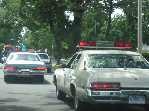 1991 Ford LTD Crown Victoria - cream sedan with a red rooftop lightbar drives alongside State Police cruisers on a tree-lined two-lane road.