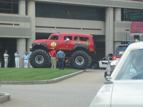 1991 Ford LTD Crown Victoria - red Extinguisher monster truck sitting on oversized tires is parked beside a concrete parking structure as people look on.