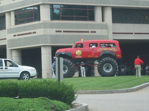 1991 Ford LTD Crown Victoria - red Extinguisher monster truck on giant tires stands near a concrete building beside a white hospital van as a crowd watches.