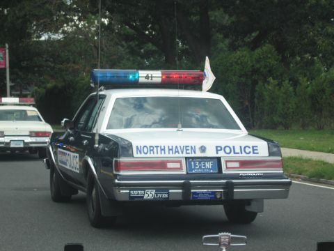 1991 Ford LTD Crown Victoria - black-and-white North Haven Police Plymouth sedan with rooftop lightbar drives down a street ahead of other cruisers.
