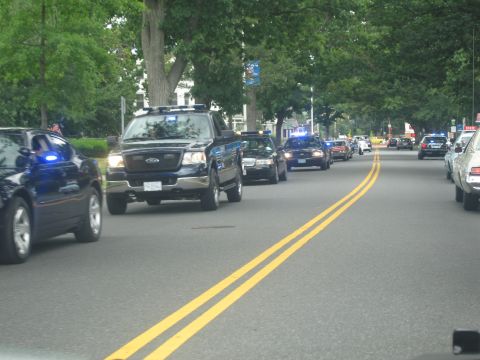 1991 Ford LTD Crown Victoria - dark Ford pickup and several police cruisers with blue lights travel along a tree-lined residential street in a parade.