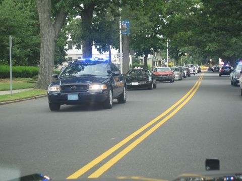 1991 Ford LTD Crown Victoria - dark Ford Crown Victoria police cruiser leads a line of vehicles down a tree-lined street with blue lights flashing behind.