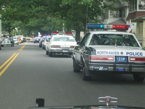 1991 Ford LTD Crown Victoria - black-and-white North Haven Police Plymouth cruiser drives in a procession of cruisers and old cars down a residential street.