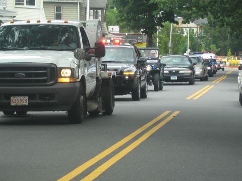 1991 Ford LTD Crown Victoria - white Ford Super Duty pickup with amber lights leads a tow truck and police vehicles down a residential street in a parade.