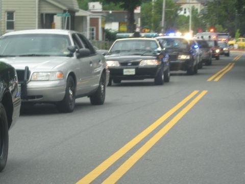 1991 Ford LTD Crown Victoria - silver Ford sedan and a column of dark police cruisers with emergency lights stretch down a residential street.