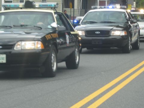 1991 Ford LTD Crown Victoria - black State Trooper Ford Mustang sits beside a black Ford police cruiser on a parade route.