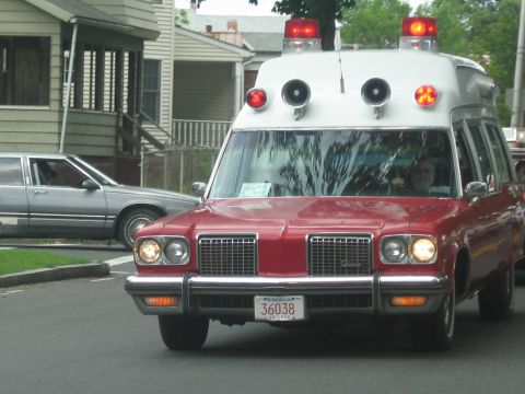 1991 Ford LTD Crown Victoria - red-and-white vintage ambulance with twin roof beacons drives through a quiet residential neighborhood.