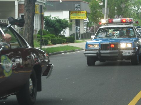 1991 Ford LTD Crown Victoria - blue police sedan with a full light bar follows a maroon cruiser past storefronts on a town street.