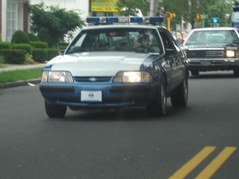 1991 Ford LTD Crown Victoria - blue-and-white State Police Ford Mustang with a Trooper-labeled light bar drives down a town road.