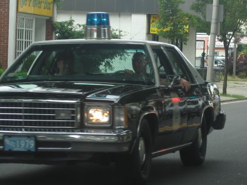 1991 Ford LTD Crown Victoria - black vintage police sedan topped with a single blue rotating beacon cruises past storefronts.