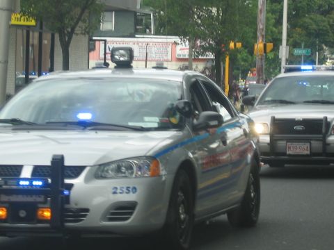 1991 Ford LTD Crown Victoria - silver police sedan marked 2550 with blue emergency lights drives alongside another cruiser on an urban street.