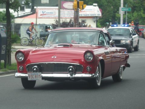1991 Ford LTD Crown Victoria - red mid-1950s Ford Thunderbird with chrome bumper and round headlights drives down a street ahead of a police car.