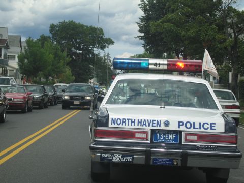 1991 Ford LTD Crown Victoria - rear of a North Haven Police sedan with a lit roof bar leads a line of vehicles down a residential street.