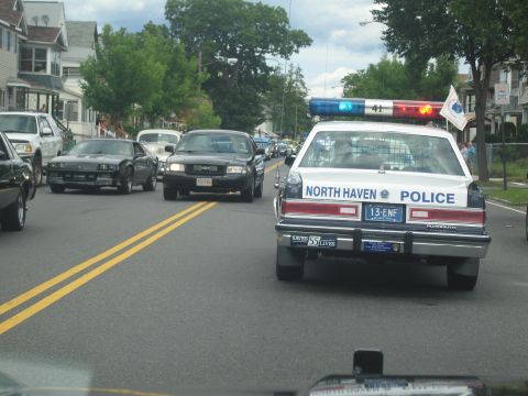 1991 Ford LTD Crown Victoria - North Haven Police cruiser with flashing roof lights leads a procession of oncoming cars down a tree-lined road.
