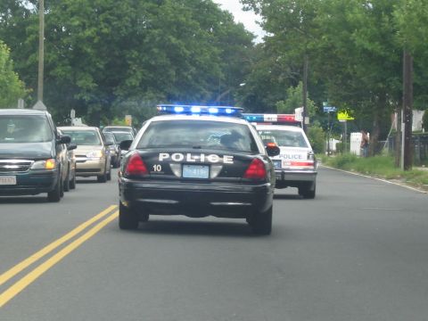1991 Ford LTD Crown Victoria - black police cruiser marked POLICE 10 with flashing blue lights leads other vehicles down a residential road.
