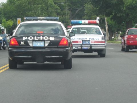 1991 Ford LTD Crown Victoria - black police cruiser marked POLICE 10 drives beside a white North Haven Police sedan with lit beacons on a road.