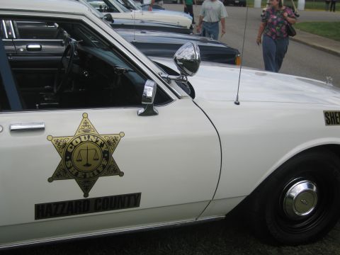 1991 Ford LTD Crown Victoria - Close-up of a white Hazzard County Sheriff car showing the gold star door decal, spotlight, and an open driver door.