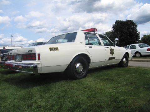 1991 Ford LTD Crown Victoria - Rear quarter view of a white Sheriff sedan with a red roof light bar and gold star decal parked on grass.