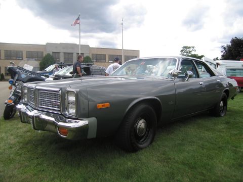 1991 Ford LTD Crown Victoria - Front-three-quarter view of a gray late-1970s Dodge four-door police sedan on grass, with a police motorcycle and parked cars behind it.