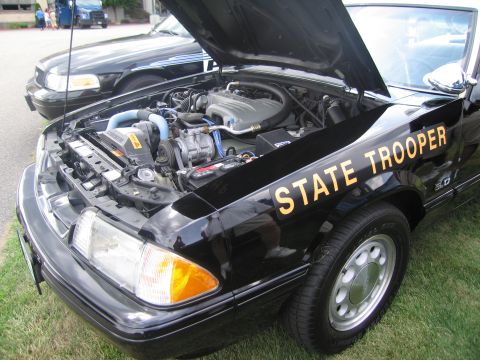 1991 Ford LTD Crown Victoria - Close view of the open engine bay of a black Ford Mustang State Trooper car, with 5.0 fender badge and yellow 'STATE TROOPER' lettering on…