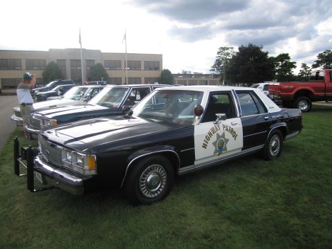 1991 Ford LTD Crown Victoria - Black-and-white Ford LTD Crown Victoria Highway Patrol sedan with a star door shield, parked in a row of cruisers in front of a large build…