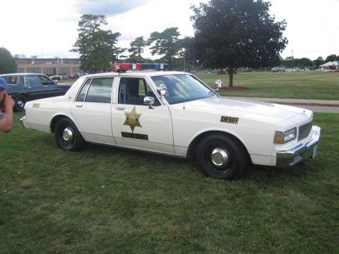 1991 Ford LTD Crown Victoria - Side view of a white Chevrolet Caprice sheriff cruiser with a gold star door decal, 'SHERIFF' lettering and roof lightbar, parked on grass…