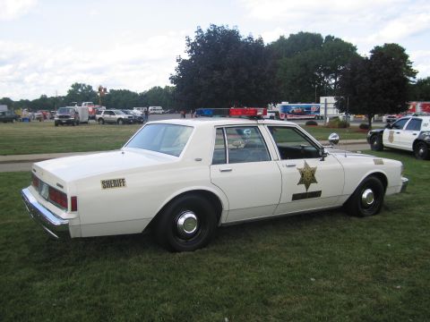 1991 Ford LTD Crown Victoria - Rear-three-quarter view of a white Chevrolet Caprice sheriff cruiser with a gold star door decal and roof lightbar, parked on grass near em…