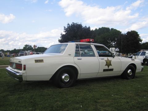 1991 Ford LTD Crown Victoria - Rear-three-quarter view of a white Chevrolet Caprice sheriff cruiser with gold star door decal, 'SHERIFF' lettering and roof lightbar, park…