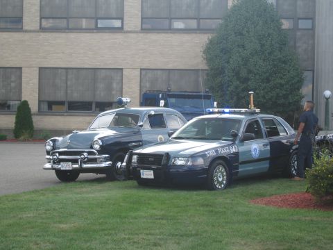 1991 Ford LTD Crown Victoria - vintage black police sedan parked beside a modern Ford Crown Victoria 'State Police 947' cruiser on grass in front of a building, with an o…