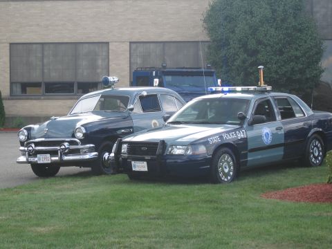 1991 Ford LTD Crown Victoria - black 1951 Ford police sedan parked next to a modern Ford Crown Victoria 'State Police 947' cruiser on grass in front of a building.