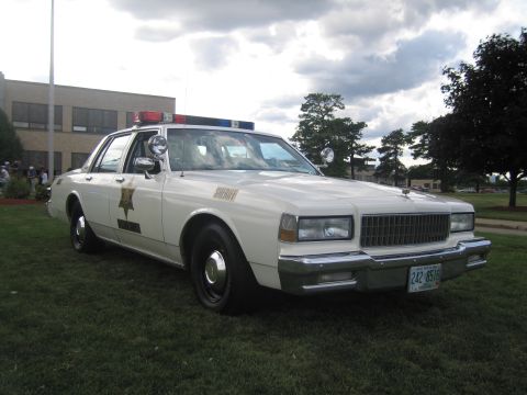 1991 Ford LTD Crown Victoria - Front-three-quarter view of a white Chevrolet Caprice sheriff cruiser with a gold star door decal, spotlight and roof lightbar, parked on g…