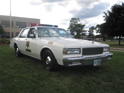 1991 Ford LTD Crown Victoria - Front-three-quarter view of a white Chevrolet Caprice sheriff cruiser with gold star door decal, 'SHERIFF' lettering, spotlight and roof li…
