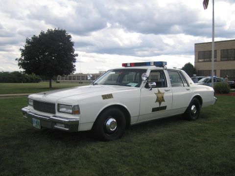 1991 Ford LTD Crown Victoria - Front-quarter view of a white Chevrolet Caprice sheriff cruiser with a gold star door decal, spotlight and roof lightbar, parked on grass n…