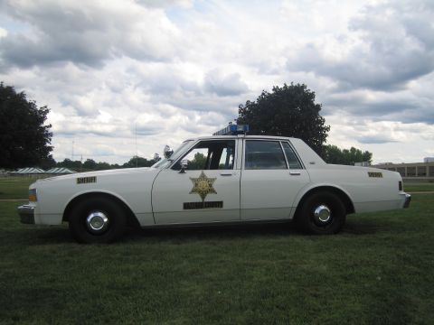 1991 Ford LTD Crown Victoria - Driver-side view of a white Chevrolet Caprice sheriff's car with gold star insignia and roof light bar on a grassy lawn.