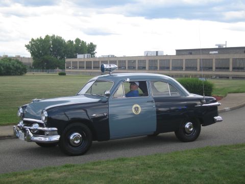 1991 Ford LTD Crown Victoria - restored early-1950s two-tone blue Ford police sedan with a roof-mounted spotlight and state seal on the door, driven on pavement.
