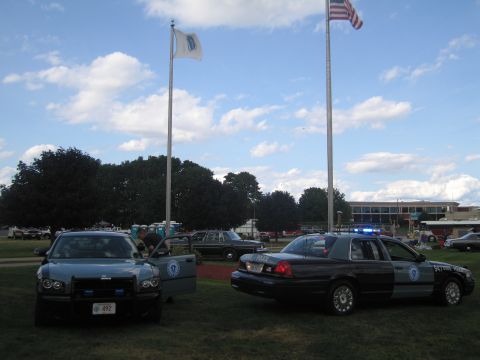 1991 Ford LTD Crown Victoria - Dodge Charger and a State Police cruiser parked on grass under an American flag and a white flag on tall poles.