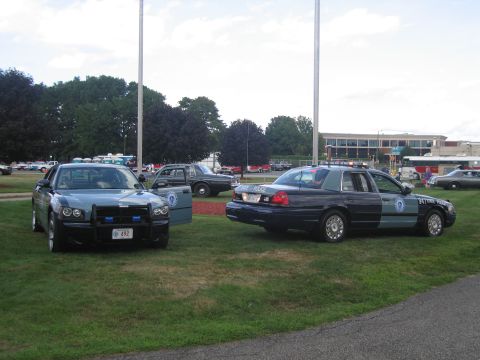 1991 Ford LTD Crown Victoria - police Dodge Charger and a State Police Crown Victoria parked on grass with other display vehicles in the background.