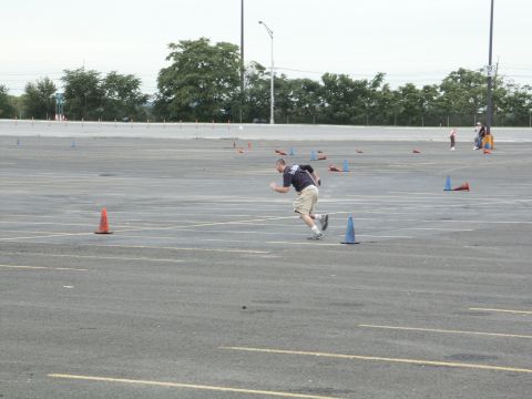 2001 Mercury Grand Marquis - A participant navigates a cone course in a large asphalt lot, with trees and distant figures visible in the background.