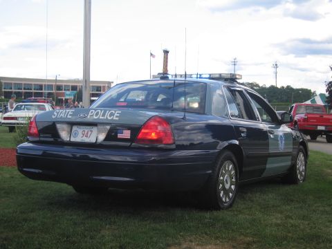 1991 Ford LTD Crown Victoria - Rear view of a dark State Police Ford Crown Victoria marked 947 with a flag decal and roof light bar, parked on grass.
