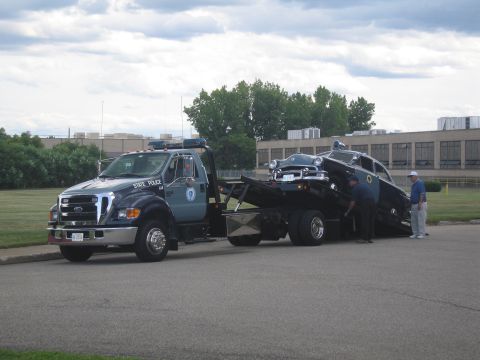 1991 Ford LTD Crown Victoria - State Police Ford flatbed tow truck loading a dark vintage police cruiser, with two men standing beside it on pavement.