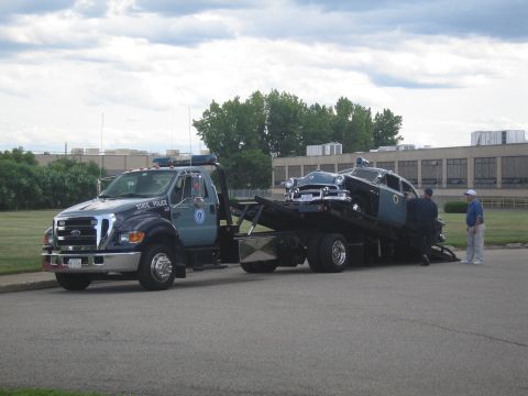 1991 Ford LTD Crown Victoria - State Police Ford flatbed tow truck hauling a dark vintage police car, with bystanders watching on a parking lot.