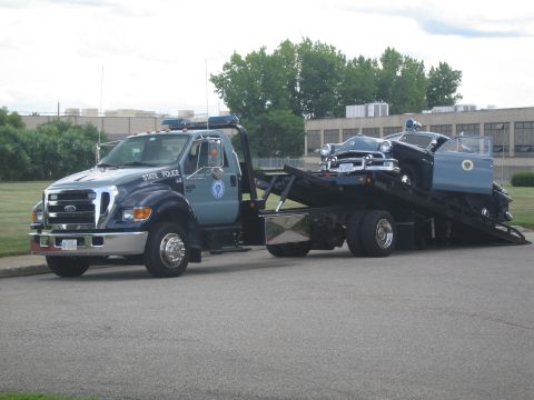 1991 Ford LTD Crown Victoria - blue-and-gray State Police Ford flatbed tow truck with a vintage police cruiser tilted on its inclined bed, on pavement.