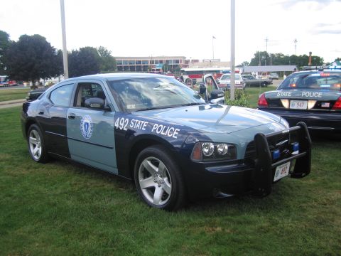 1991 Ford LTD Crown Victoria - blue-and-gray State Police Dodge Charger numbered 492 with push bar on grass, with cruisers and buildings behind.