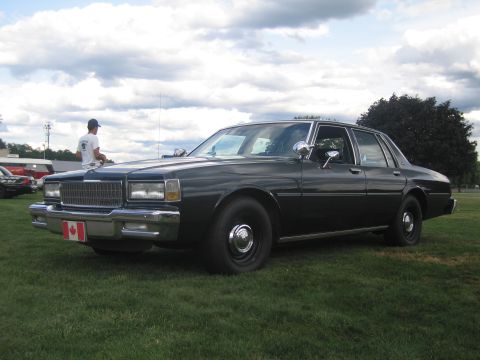 1991 Ford LTD Crown Victoria - black Chevrolet Caprice sedan with a Canadian flag front plate and roof spotlight, parked on grass under a cloudy sky.
