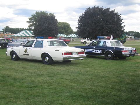 1991 Ford LTD Crown Victoria - white sheriff's patrol sedan and a dark North Haven Police cruiser displayed side by side on a grassy field at a car show.