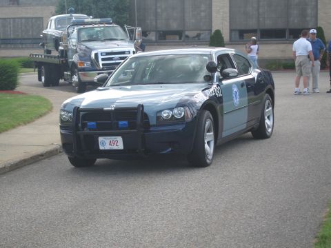 1991 Ford LTD Crown Victoria - dark blue police sedan fitted with a front push bar rolls along a road past a flatbed tow truck and onlookers.